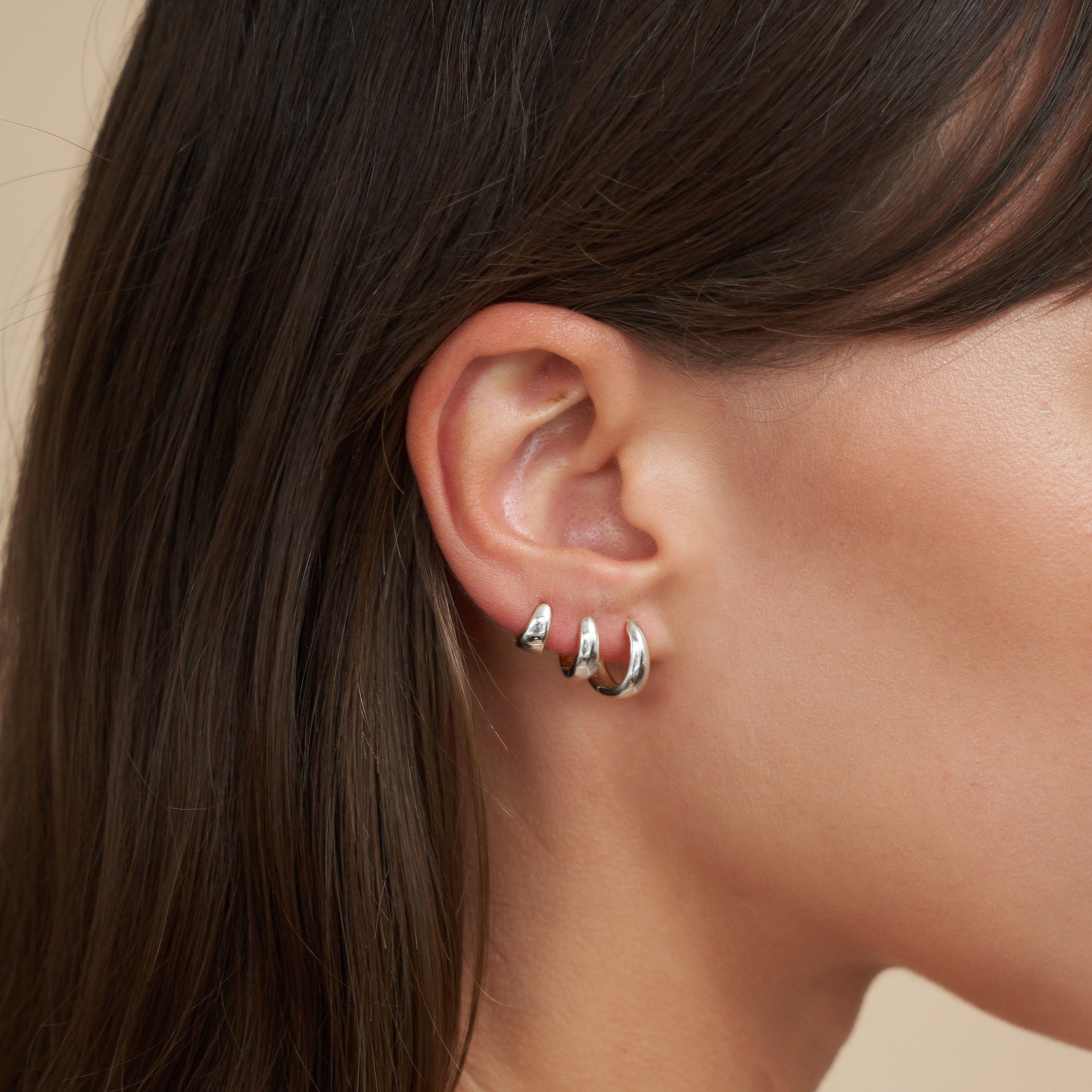 Close-up of an ear wearing a silver hoop earring with dark hair and beige background