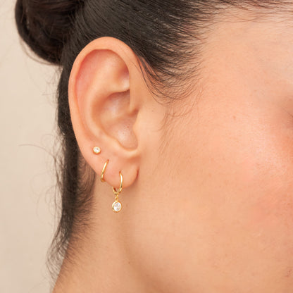 Close-up of an ear wearing gold hoop earrings with small gemstones on a neutral background