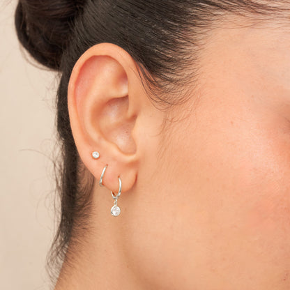 Close-up of an ear wearing two silver hoop earrings with small round stones on a beige background