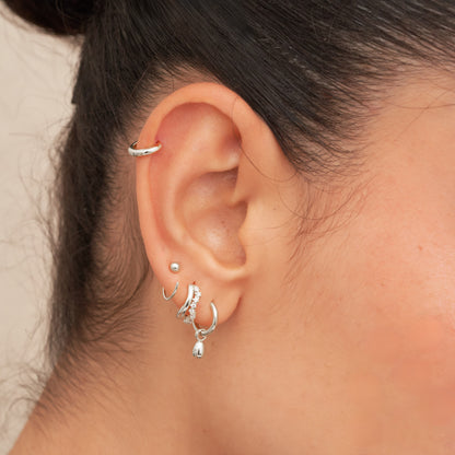 Close-up of an ear with silver earrings and a ring, against a neutral background.
