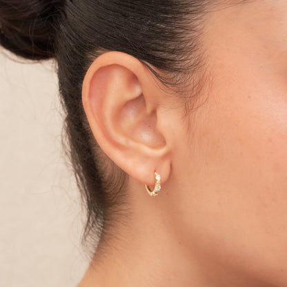 Close-up of a person wearing a gold earring with a small gemstone on a neutral background