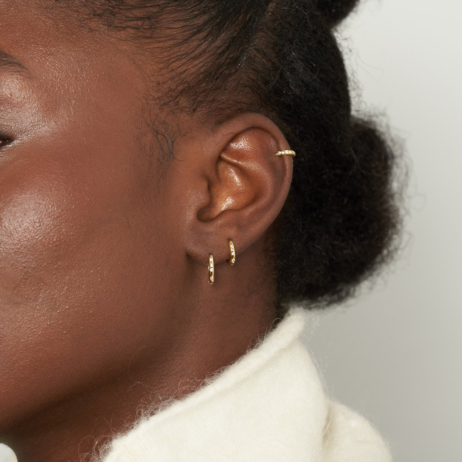 Close-up of a person's ear with gold hoop earrings on a neutral background