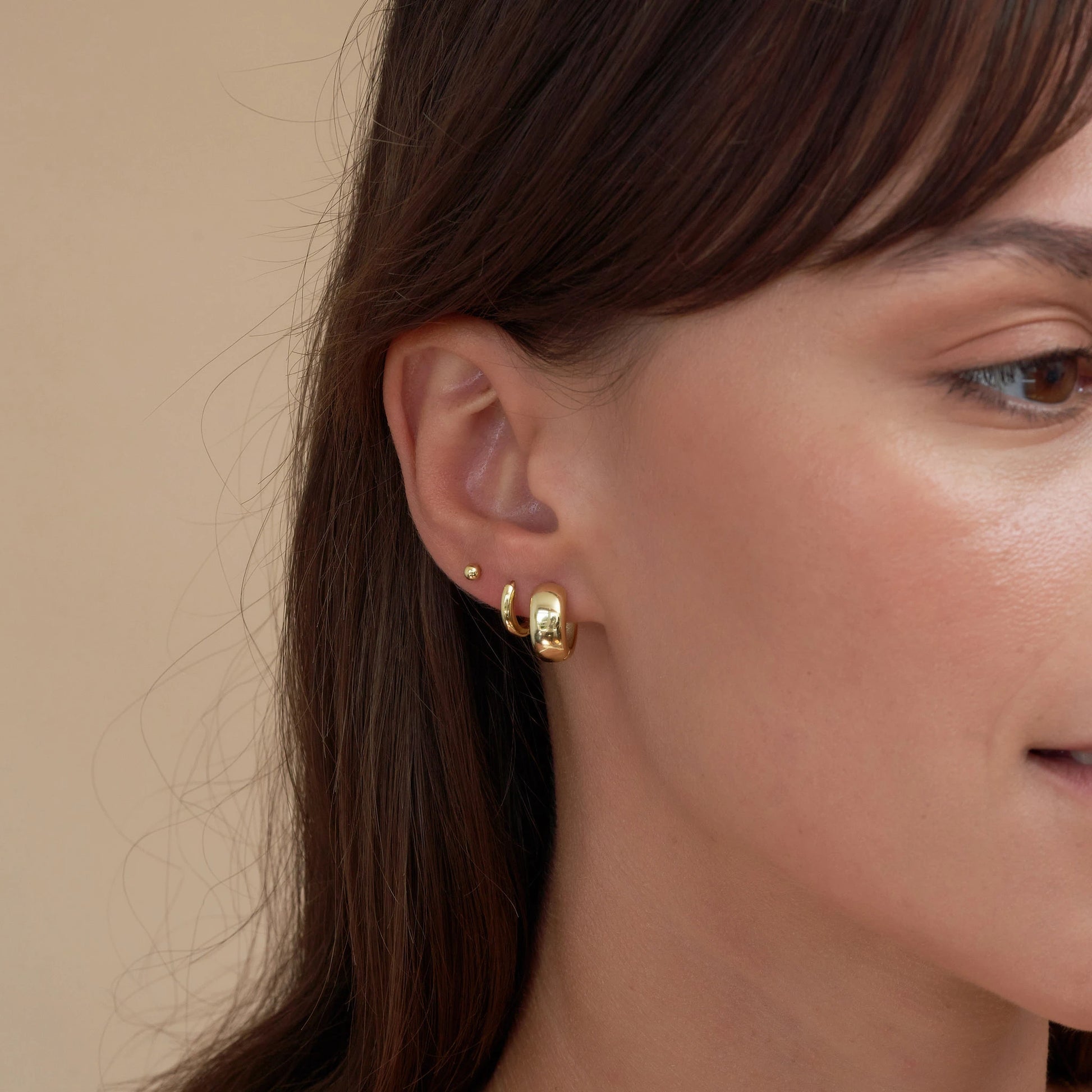Close-up of a person wearing gold hoop earrings against a neutral background