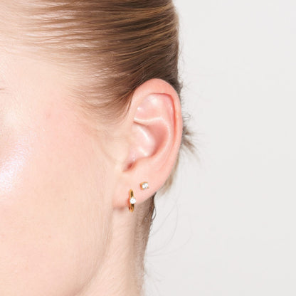 Close-up of an ear wearing a gold earring with a clear gemstone on a neutral background