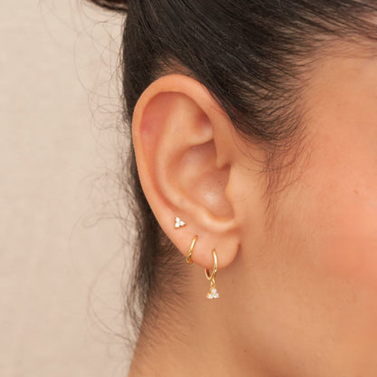 Close-up of an ear wearing gold hoop earrings with small heart and diamond-shaped charms on a neutral background.