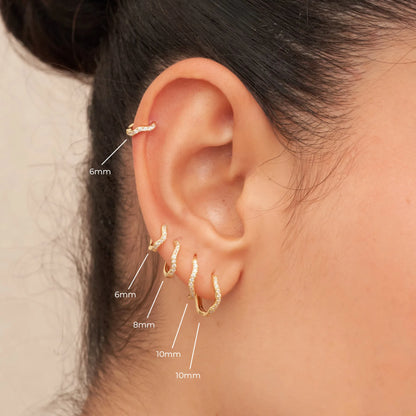 Close-up of an ear with gold hoop earrings labeled by size, on a neutral background.